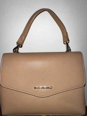Beige handväska från Valentino - Snygg beige handväska från Valentino i stilren design. Väskan har ett avtagbart axelband i silverfärgad kedja och ett kortare handtag i matchande läder. Tillverkad i slätt, strukturerat läder med guldfärgad logga framtill.