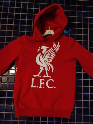 Röd LFC hoodie med tryck - Snygg röd hoodie med stort vitt Liverpool FC-tryck på bröstet och klassisk huva. Perfekt för dig som vill visa ditt stöd för laget. Mjuk och skön bomullsblandning, långärmad och med ribbade muddar.