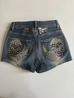 Blå jeansshorts med döskalletryck - Snygga blå jeansshorts med coola döskalletryck och vingar på bakfickorna samt döskallemönster i linningen. Shorts i klassisk denim med slitningar och nitar för extra edge. Perfekta för dig som vill sticka ut i sommar! paketpris om du köper mer
