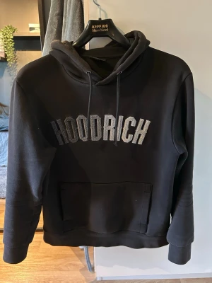 Svart hoodie från Hoodrich - Svart hoodie från Hoodrich med stor broderad logga i grått på bröstet. Klassisk modell med huva, dragsnören och stor magficka framtill. Perfekt för en avslappnad streetwear-look. Mjuk och skön bomullsblandning.