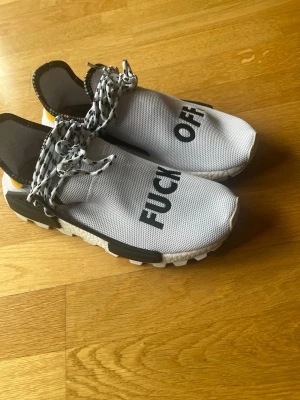 Vita Pharrell x Adidas Hu NMD sneakers - Unika vita Pharrell x Adidas Hu NMD sneakers med svart text på ovandelen. Skorna har svart och vitmönstrade snören, svart sula och detaljer samt färgglada inslag på insidan av kragen. Ovandelen är i mesh-material för skön komfort.