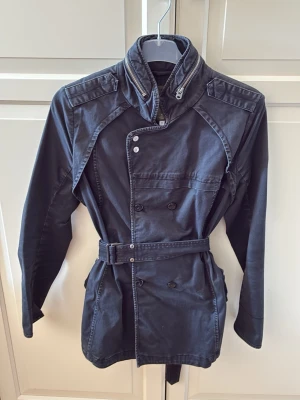 Svart trenchcoat med bälte och hög krage från G-Star RAW - Stilren svart trenchcoat med dubbelknäppning, hög krage med dragkedjedetalj och axelklaffar. Jackan har ett avtagbart bälte i midjan och flera fickor framtill. Storlek M, men passar S bättre. Jackan har blivit lite uttvättad, så att den svarta färgen har försvunnit lite. Annars jackan i fint skick. 
