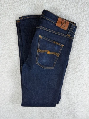 Nudie jeans Lean Dean (Dry 16 Dips) W33 L34 - Snygga mörkblå jeans från Nudie med klassiska orange sömmar och läderpatch bak i midjan. Jeansen har fem fickor och en smal passform som sitter tight hela vägen ner. Perfekta för dig som gillar stilrena och moderna jeans.