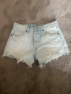 Ljusblå denimshorts med fransig kant - Snygga ljusblå denimshorts med hög midja och fransig, rå kant nertill. Klassisk femficksmodell med knapp  framtill. Perfekta för sommaren och ger en avslappnad vibe.