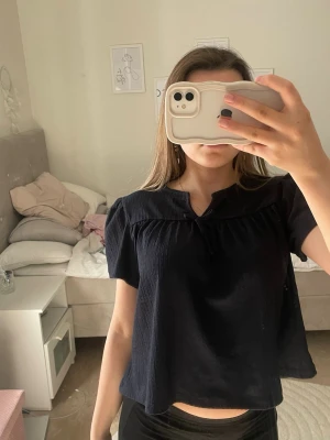 Svart blus med kort ärm - Svart blus med korta puffärmar och lös passform. Blusen har en liten v-ringning med dekorativ detalj vid halsen och är tillverkad i ett lätt, luftigt tyg. Perfekt för en stilren och enkel look.