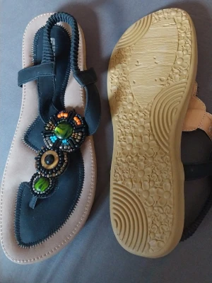 Svarta sandaler med pärldekor - Snygga svarta sandaler med elastiskt band och dekorativa pärlor i grönt, orange och blått framtill. Sulan är beige med svarta detaljer och har mönstrad undersida för bra grepp. Perfekta för en trendig och avslappnad look