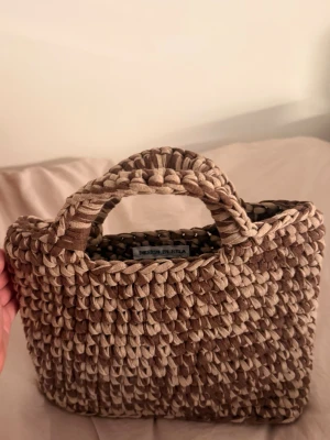 Virkad väska i bruna nyanser  - Väskan är brunbeige och har rejäla handtag. Perfekt accessoar för en trendig look och rymmer det viktigaste du behöver ha med dig. Virkad i återvunnet material. 