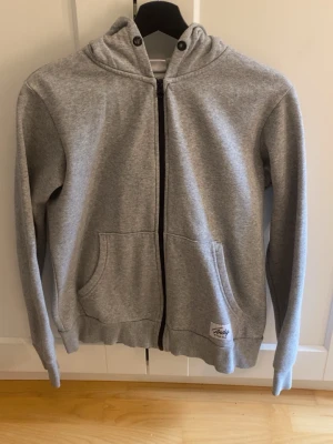 Grå zip-hoodie från Andy - Snygg grå hoodie med dragkedja. Klassisk modell med huva, två stora fickor framtill och ribbade muddar vid ärmslut och nederkant. Perfekt för en chill och avslappnad stil. Liten logga nertill på ena fickan.