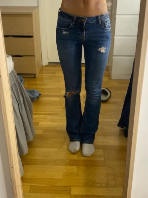 Blå slitna bootcut jeans - Säljer ett par blå bootcut jeans med slitningar och hål på låret och knät. Jeansen har klassisk femficksmodell, låg midja och dragkedja med knapp. Perfekta för en avslappnad och trendig look.