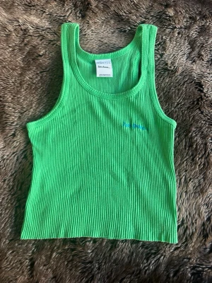 Grön ribbad linnetopp  - Snygg grön ribbad linnetopp med broderad blå text på bröstet. Toppen är ärmlös och har en tight passform, perfekt att styla med jeans eller kjol. Materialet är mjukt och stretchigt för extra komfort.