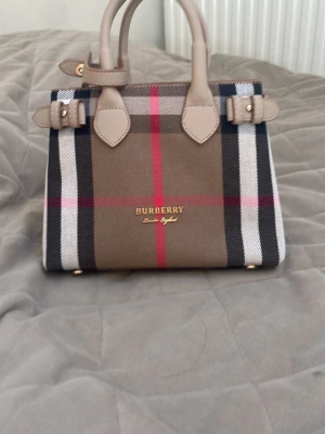 Burberry rutig minihandväska - Snygg minihandväska från Burberry i klassiskt rutigt mönster med beige, svart, vitt och rött. Väskan har två handtag och detaljer i ljusbeige läder samt guldfärgade metalldetaljer. Perfekt för dig som gillar lyxiga accessoarer med ikonisk design.