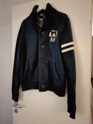 Abercrombie & Fitch Marinblå - Marinblå cardigan jacka från Abercrombie & Fitch med stora knappar, hög ribbad krage och två vita ränder på ena ärmen. Tryck med A&F 92 på bröstet och två fickor framtill. Sportig och avslappnad stil i mjukt bomullsmaterial.