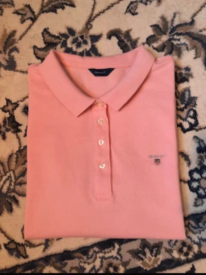 Rosa piké t-shirt från GANT - Snygg rosa pikétröja från GANT med klassisk krage och tre knappar framtill. Tröjan har korta ärmar och diskret broderad logga på bröstet. Perfekt för en stilren och avslappnad look. Mått på bilder, hör av er vid funderingar. Pris kan diskuteras vid snabb affär. Etiketten visar strl L men sitter som s/m