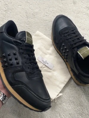 Svarta Valentino sneakers med nitar - Snygga svarta sneakers från Valentino med coola svarta nitar på sidorna och beige sula. Skorna är gjorda i en mix av läder och mocka, har svarta skosnören och en logga på plösen. Perfekta för dig som vill sticka ut med en exklusiv touch.