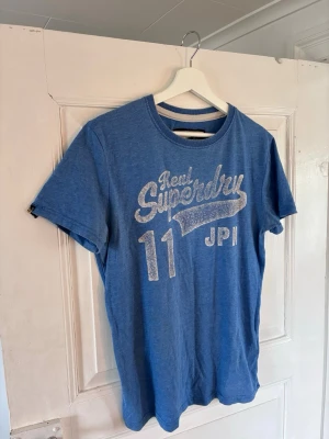 Blå Superdry t-shirt med tryck - Snygg blå t-shirt från Superdry med stort vitt tryck framtill och rund hals. Modellen är klassisk med korta ärmar och normal passform. Perfekt för en avslappnad och cool stil. Materialet är mjuk bomull som känns skönt mot huden.