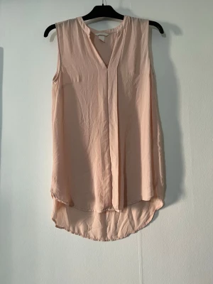 Ljusrosa ärmlös blus med v-ringning - Säljer en ljusrosa ärmlös blus med v-ringning och längre rundad nederkant. Blusen har en lös och flowy passform, perfekt för en avslappnad och stilren look. Tillverkad i ett tunt och lätt material som känns svalt mot huden.