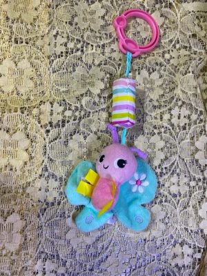 Färgglad fjäril musik babyleksak med klämma - Söt babyleksak i form av en fjäril med turkosa vingar, rosa kropp och lila antenner. Har en rosa plastklämma för enkel upphängning och en randig cylinder i pastellfärger. Dekorerad med en liten blomma och prassliga detaljer. Helt ny 