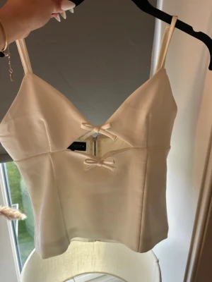 Korsettopp Zara - Snygg champagnefärgad korsettopp i satin med smala axelband, två små rosetter framtill och v-ringad urringning. Toppen har en dragkedja i ryggen och en lätt glansig finish som ger en lyxig känsla. Perfekt för att lyfta vilken outfit som helst.