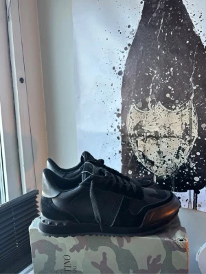 Svarta Valentino sneakers med camo - Snygga svarta sneakers från Valentino med diskret camo-mönster och nitar på sulan. Skorna har en klassisk låg siluett, svarta skosnören och detaljer i både läder och mocka. Perfekta för dig som gillar exklusiva och stilrena sneakers.