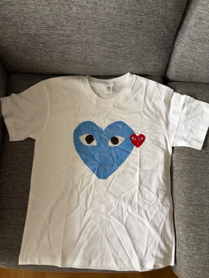 Cdg t shirt - Vit t-shirt med stort blått hjärta med ögon och ett mindre rött hjärta med ögon på bröstet. Klassisk rund halsringning och korta ärmar. Perfekt för dig som gillar grafiska tryck och vill sticka ut lite extra.