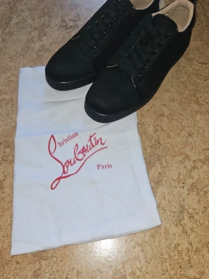Svarta Christian Louboutin sneakers - Snygga svarta sneakers från Christian Louboutin med klassisk röd sula och svarta skosnören. Skorna har en låg profil och är tillverkade i ett material som ser ut som mocka eller nubuck. Perfekta för dig som vill sticka ut med exklusiva detaljer. Helt nya, finns k storlek 44 och 43