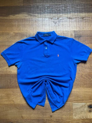 Polo Ralph Lauren blå piké S - Polo Ralph Lauren piké i blå färg med rosa logga. Storlek S och sitter snyggt fitted. Mycket bra skick utan större slitage. Perfekt till sommaren eller clean vardagsfit.
