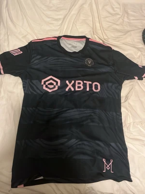 Inter Miami svart matchtröja - Snygg svart Inter Miami matchtröja från Adidas med rosa detaljer och zebramönster. Tröjan har klubbmärke på bröstet, sponsorlogga 'XBTO' i rosa och rund halsringning med rosa kant. Perfekt för fotboll eller streetwear.