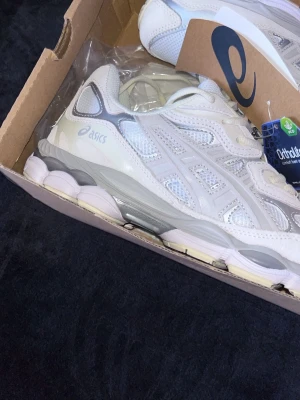Vita Asics sneakers med mesh och skinn - Säljer ett par fräscha vita Asics sneakers med detaljer i grått och silver. Skorna har en chunky sula, meshpaneler för bra ventilation och overlays i syntetiskt skinn. Klassisk Asics-logga på sidan och Ortholite-innersula för extra komfort storlek 40-41. 