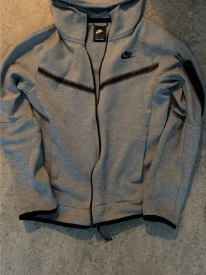 Grå Nike hoodie med svarta detaljer - Snygg grå hoodie från Nike med svarta dragkedjor och logga på bröstet. Hoodien har huva, hel dragkedja framtill och en extra ficka med dragkedja på ena ärmen. Tillverkad i mjukt material som känns bekvämt och sportigt.