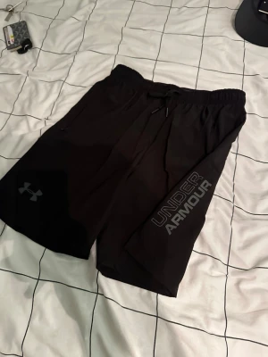 Svarta träningsshorts från Under Armour - Säljer ett par svarta träningsshorts från Under Armour med logga och text på ena benet. Shortsen har elastisk midja med snörning och är tillverkade i ett lätt och snabbtorkande material. Perfekta för gymmet eller löprundan.