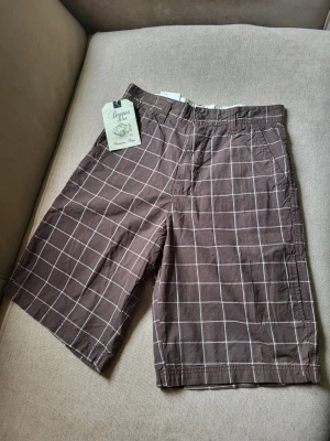Bruna rutiga shorts från Banan Blue 32 - Bruna shorts från Dynamic Style med vitt rutmönster. Klassisk passform med fickor fram och bak samt bälteshällor. Tillverkade i bomull, perfekta för varma dagar. Stängs med knapp och dragkedja. Snygg och avslappnad stil för sommaren.