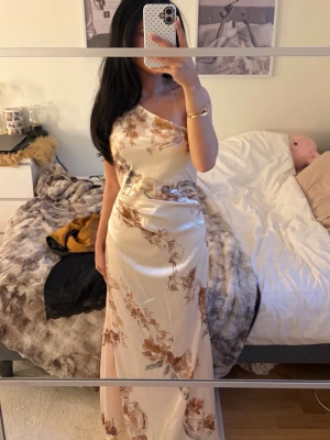 Champagnefärgad oneshoulder långklänning - Elegant långklänning i champagnefärg med bruna blommiga mönster. Klänningen har oneshoulder-design och är tillverkad i ett glansigt satinliknande material som ger en lyxig känsla. Perfekt för speciella tillfällen när du vill sticka ut.