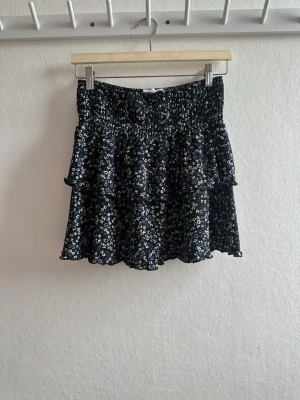 Blommig volangkjol från Primark - Supersöt kort kjol från Primark med små vita och blå blommor på svart botten. Kjolen har volanger och smock i midjan för en bekväm passform. Perfekt för dig som gillar en lekfull och trendig stil.