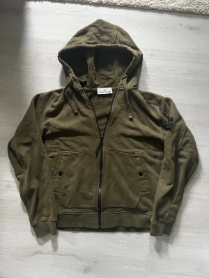 Olivgrön hoodie från Stone Island - Snygg olivgrön hoodie från Stone Island med dragkedja framtill, två stora fickor och klassisk patch med logga på vänster ärm. Tillverkad i mjuk bomull och har ribbade muddar samt justerbar huva med snören. Perfekt för en avslappnad streetwear-look.