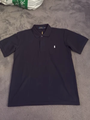 Svart pikétröja från Polo Ralph Lauren - Klassisk svart pikétröja från Polo Ralph Lauren med vit broderad logga på bröstet. Tröjan har korta ärmar, krage och knappar framtill. Perfekt för en clean och stilren look.