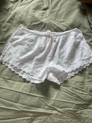 Vita spetsdetaljerade shorts - Säljer ett par vita shorts med broderade spetsdetaljer och vågig kant. Shortsen har elastisk midja med liten rosett framtill. Helt nya och oanvända med prislapp.