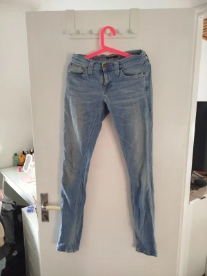 Ljusblå skinny jeans från Nudie. W27 L34 - Säljer ett par ljusblå skinny jeans från Nudie Jeans med snygga detaljer på bakfickorna. Jeansen är tillverkade i mjuk denim med stretch för extra komfort. Perfekta till en avslappnad  och snygg stil. Storleken är W27 och L34