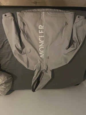 Ljusgrå vindjacka från Moncler - Snygg ljusgrå vindjacka från Moncler med stor logga framtill, två dragkedjeförsedda fickor och huva. Jackan har minimalistisk design och liten Moncler-detalj på ryggen. Perfekt för dig som vill ha en stilren och exklusiv look.