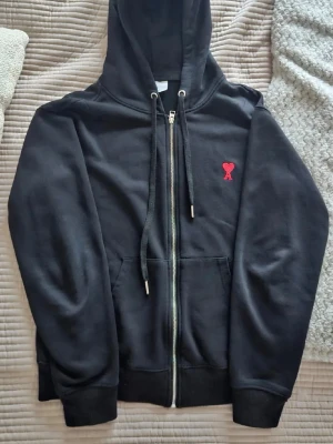 Svart hoodie med dragkedja från Ami - Svart hoodie från Ami Alexandre Mattiussi med dragkedja framtill, huva med snören och röd broderad logga på bröstet. Tillverkad i mjukt material och har fickor på sidorna. Perfekt för en avslappnad och stilren look.