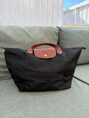 Svart Longchamp Le Pliage handväska - Klassisk svart Le Pliage handväska från Longchamp med bruna läderhandtag och lock. Väskan har guldfärgad knapp och är tillverkad i slitstark nylon med detaljer i präglat läder. Perfekt för dig som gillar stilren och tidlös design.
