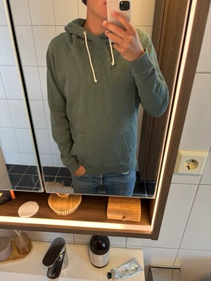 Grön hoodie från Abercrombie & Fitch - Snygg grön hoodie från Abercrombie & Fitch med klassisk känguruficka och vita dragsnören i huvan. Mjuk bomullskänsla och relaxed fit, perfekt för chill dagar. Enkel och stilren design utan tryck eller logga på framsidan.
