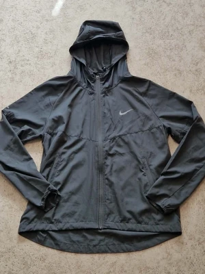 Svart Nike vindjacka med huva - Snygg svart vindjacka från Nike med hel dragkedja och huva. Jackan har diskret Nike-logga på bröstet, två sidofickor och elastiska muddar vid ärmsluten. Tillverkad i lätt och tunt material som passar perfekt för blåsiga dagar.