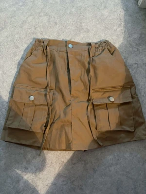 Beige utility skort med fickor - Snygg beige utility skort med stora cargofickor framtill och silverfärgade knappar. Skortsen har elastisk midja och dragsko för skön passform. Tillverkad i ett slitstarkt bomullsliknande material. Perfekt för dig som gillar streetwear och praktiska detaljer. Köpt utomlands för ca ~150-200kr. Okänt märke. Sitter tajt. Aldrig använd då den var för liten. Rekommenderad storlek S-M. Sista bilderna är en REFERENS inte riktiga kjolen. INBYGGDA SHORTS INUTI KJOLEN