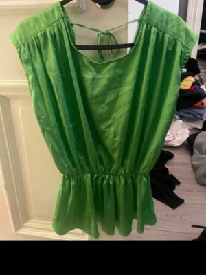 Grön ärmlös kort klänning med knyt - Snygg grön kortklänning i glansigt material med resår i midjan och volang nedtill. Klänningen är ärmlös och har ett dekorativt knyt i nacken. Perfekt för sommarens alla tillfällen.