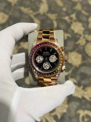 Seiko mod rainbow Daytona  - Snygg Seiko klocka i guld med länkarmband och svart urtavla. Klockan har regnbågsfärgade stenar runt boetten och tre mindre urtavlor. Extra detaljer med gnistrande stenar på sidorna av boetten. Perfekt accessoar för dig som gillar att sticka ut. Har använt mig av en VK63 som går på batteri. 