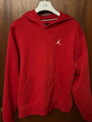 Röd Jordan hoodie  - Snygg röd hoodie från Jordan med klassisk huva och känguruficka framtill. Diskret vitt Jumpman-logo broderat på bröstet. Tillverkad i mjukt material som känns skönt mot huden. Perfekt för dig som gillar streetwear och sportig stil.