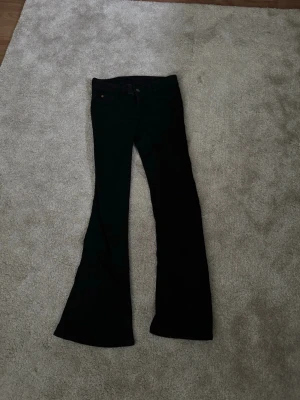 Svarta bootcut jeans  - Säljer ett par svarta bootcut jeans med utsvängda ben och en lila patch bak i midjan. Klassisk femficksmodell och låg midja. Perfekta för dig som gillar retrostil och vill ha ett par jeans som sticker ut lite extra. Storlek 26/30