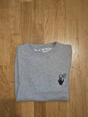 Grå sweatshirt från Off-White - Säljer en grå sweatshirt från Off-White med svart tryck på bröstet och röda samt svarta detaljer längs ärmarna. Tröjan har rund halsringning och är långärmad. Perfekt för dig som gillar streetwear och vill sticka ut lite extra.