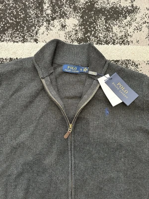 Grå ziptröja Polo Ralph Lauren - Mörkgrå ziptröja från Polo Ralph Lauren med hög krage och dragkedja hela vägen. Tröjan har två fickor framtill, ribbade muddar och en liten blå broderad logga på bröstet. Materialet är mjukt och stickat för en clean och klassisk stil. OBS: LITEN I STORLEKEN 