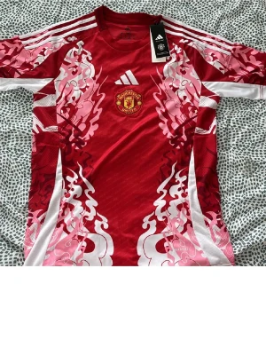 Manchester United Adidas matchtröja - Säljer en röd Manchester United matchtröja från Adidas med vitt och rosa grafiskt mönster. Tröjan har klubbmärket på bröstet, vita Adidas-ränder på axlarna och är tillverkad i ett lätt, ventilerande material. Perfekt för fotboll eller träning.